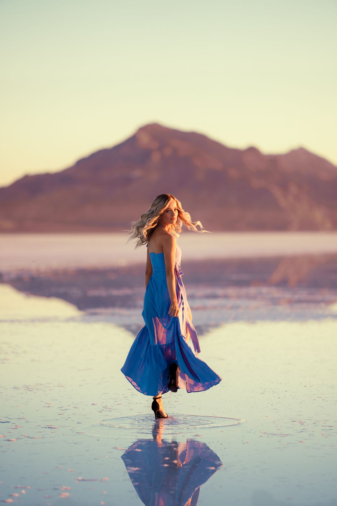 Figure in flowing blue dress twirls on reflective salt flats at sunset with mountains in background.