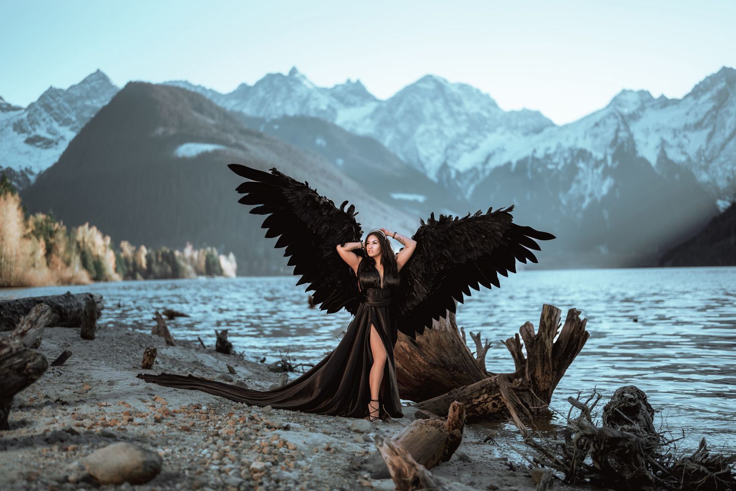 Dark figure with black wings poses dramatically by a mountain lake during sunset with snowy peaks in background.