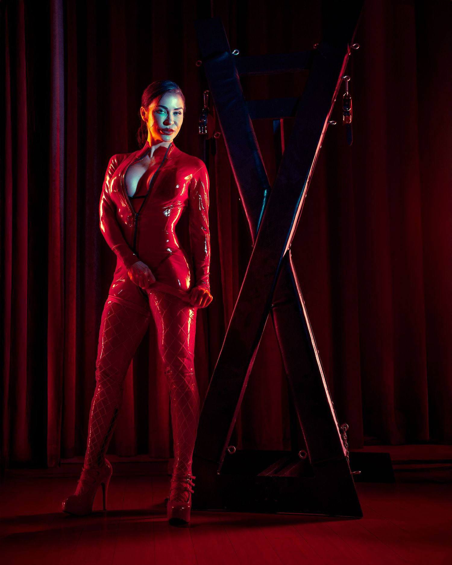 Model in red latex outfit poses dramatically next to large X-frame in moody dark setting.