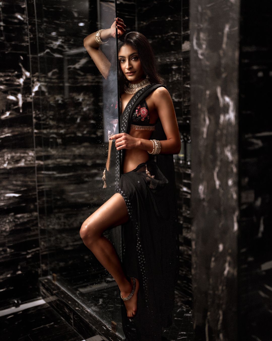 Woman in black sari strikes elegant pose against dark marble wall with dramatic lighting.