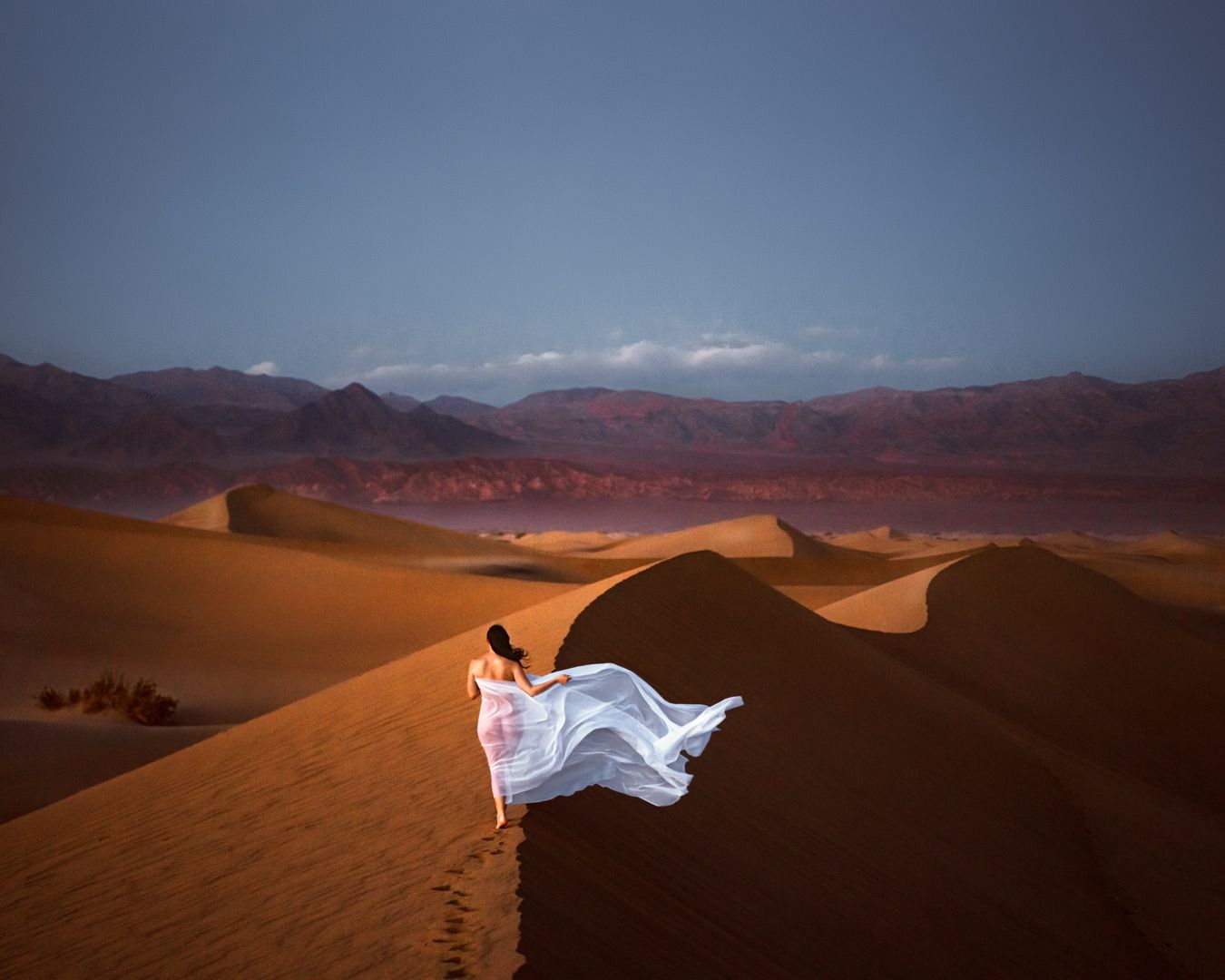 Figure in flowing white dress stands on sand dune against mountainous desert landscape.