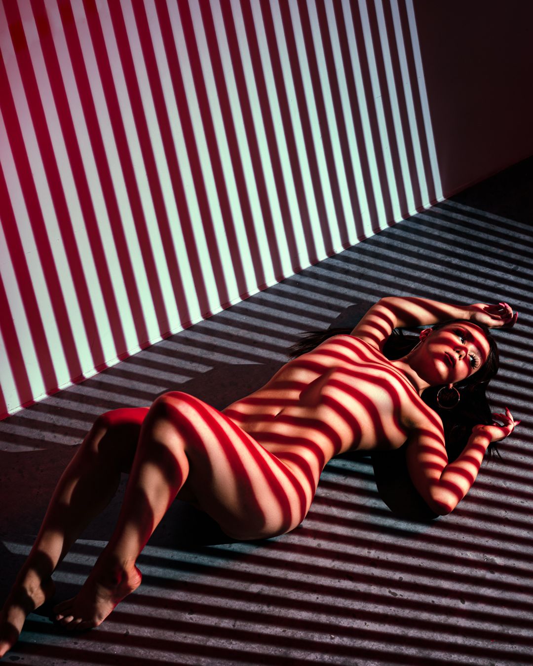 Artistic nude study with striped light patterns creating geometric shadows.
