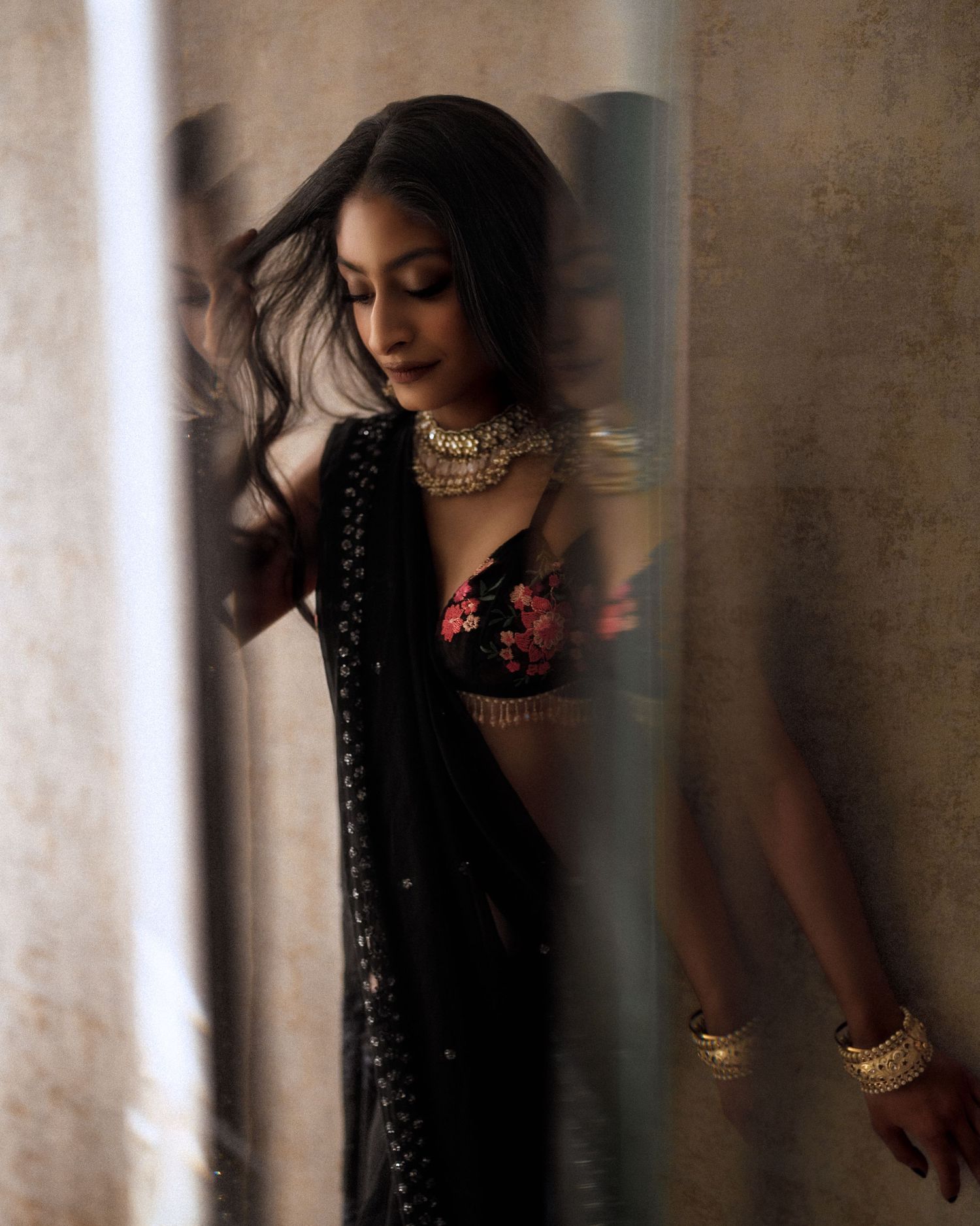 Fashion portrait of woman in black saree and ornate jewelry against textured wall.