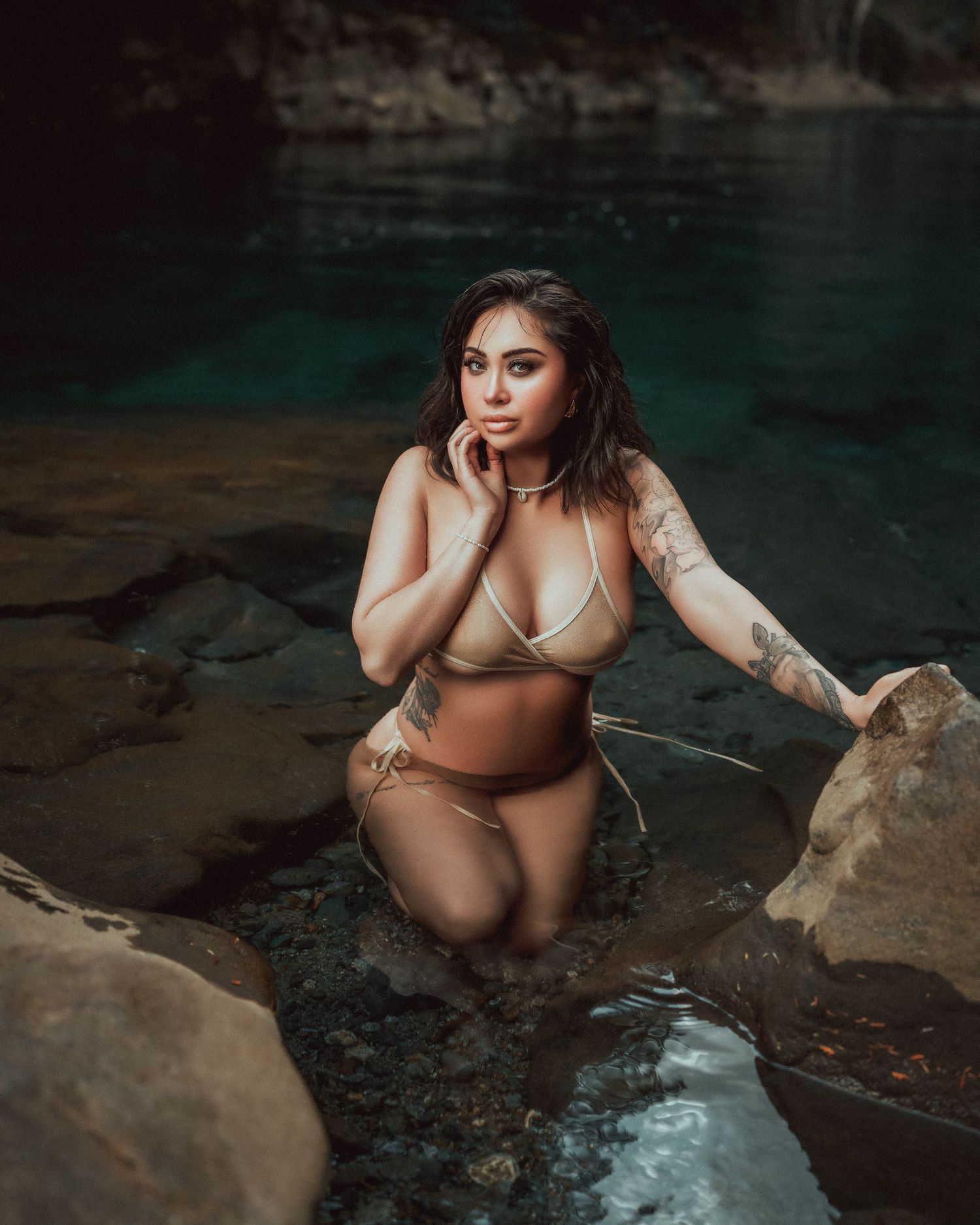 Moody outdoor boudoir photography at a rocky waterfront location with teal-colored water in background.
