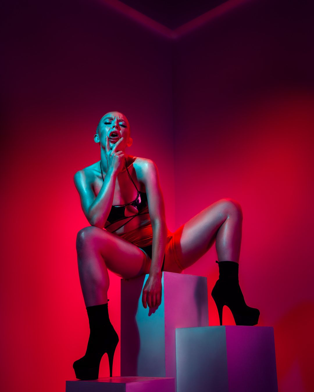 Dramatic red and blue lit artistic portrait with figure posed on geometric blocks wearing boots.
