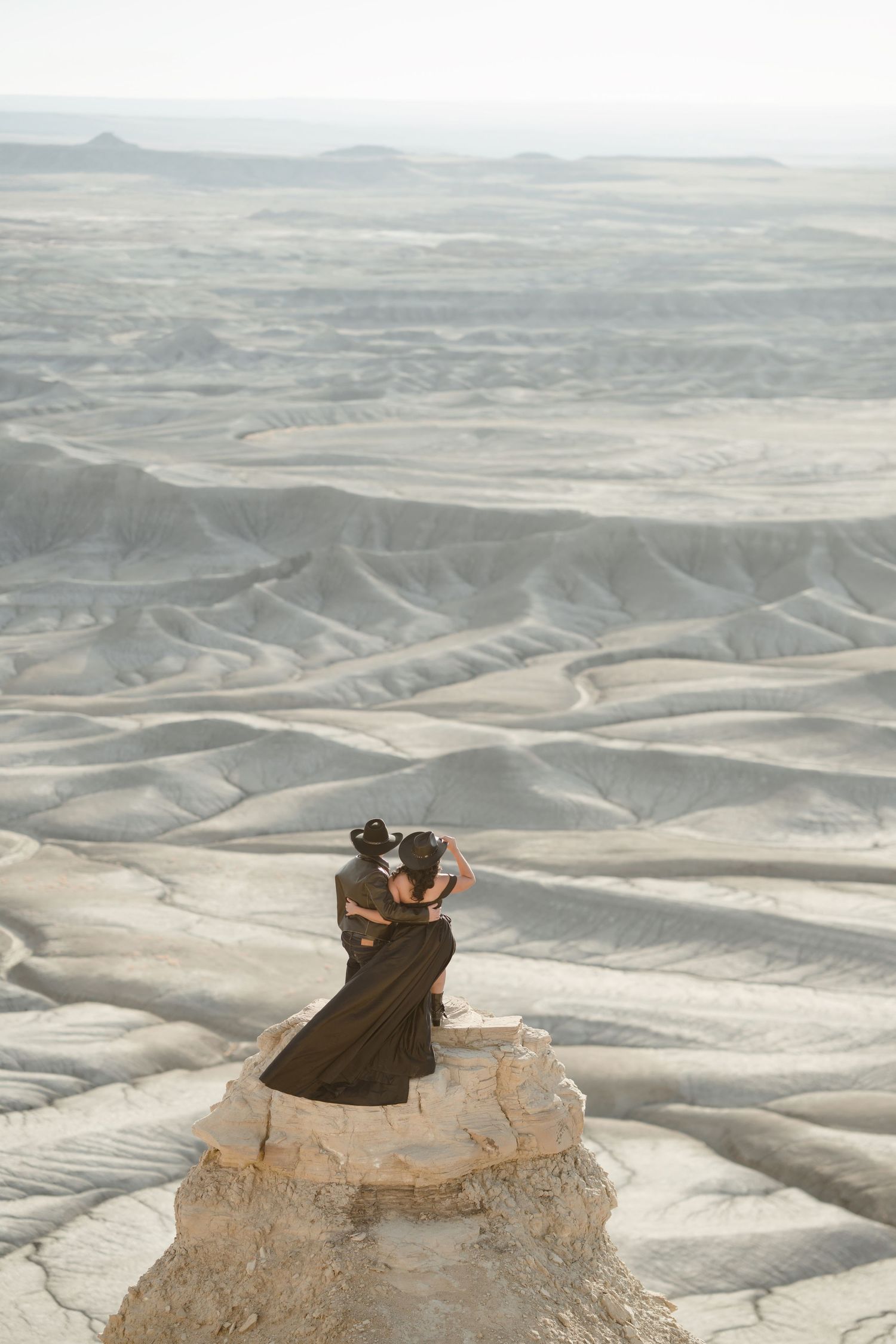 Couple standing atop dramatic desert mountain peak overlooking vast eroded valley landscape.