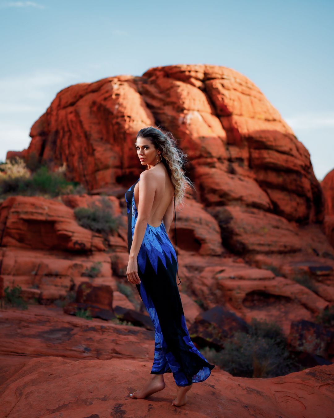 Figure in flowing blue dress poses against dramatic red rock formations in desert setting.