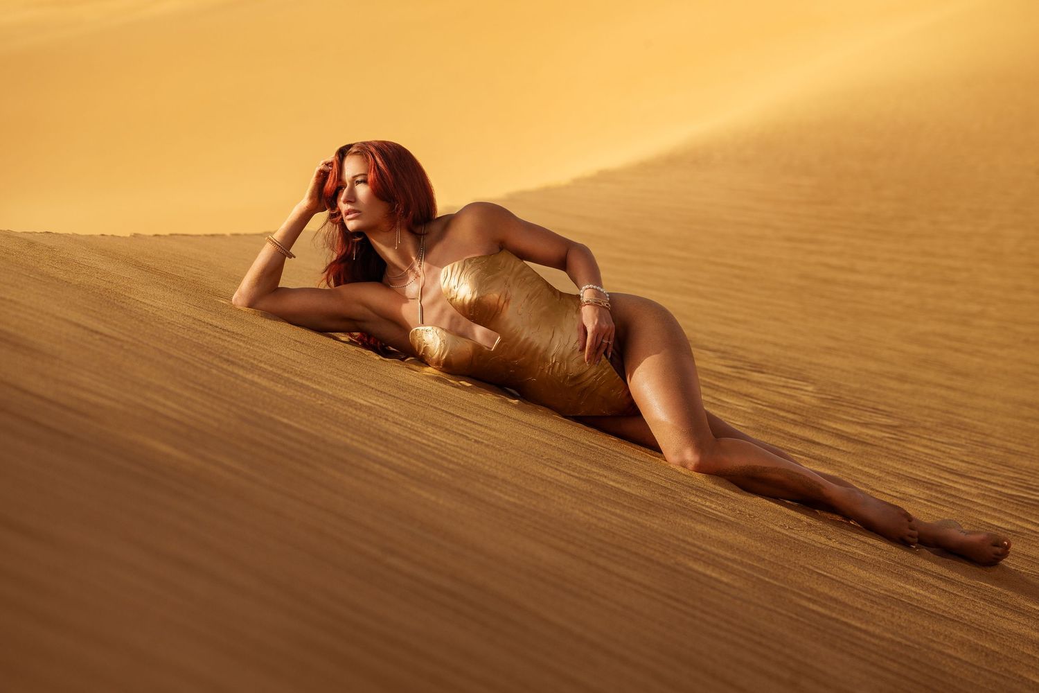 Model in golden swimsuit poses elegantly on sand dunes during golden hour sunset in desert setting.