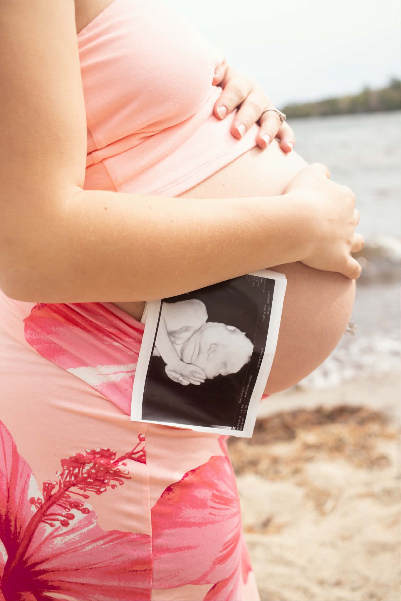 Pregnant mother wearing a bathing suit holding her belly and showing a 3-D ultrasound photo at the beach.