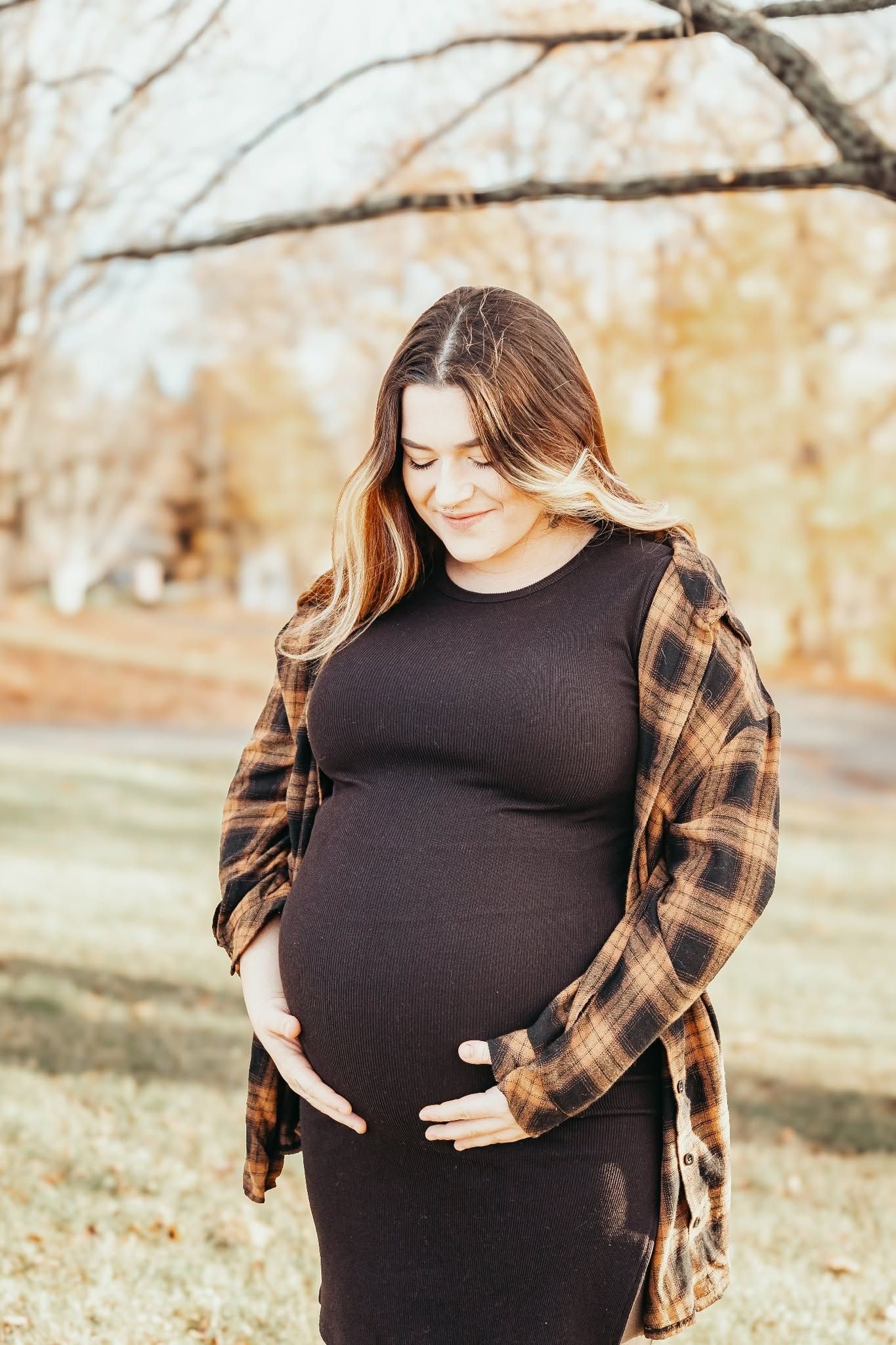 Pregnant mother holding her belly wearing a black dress and flannel in the fall woods.