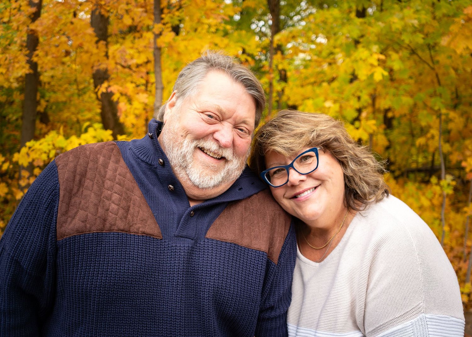 A couple smiles warmly together outdoors against a backdrop of vibrant yellow autumn foliage.