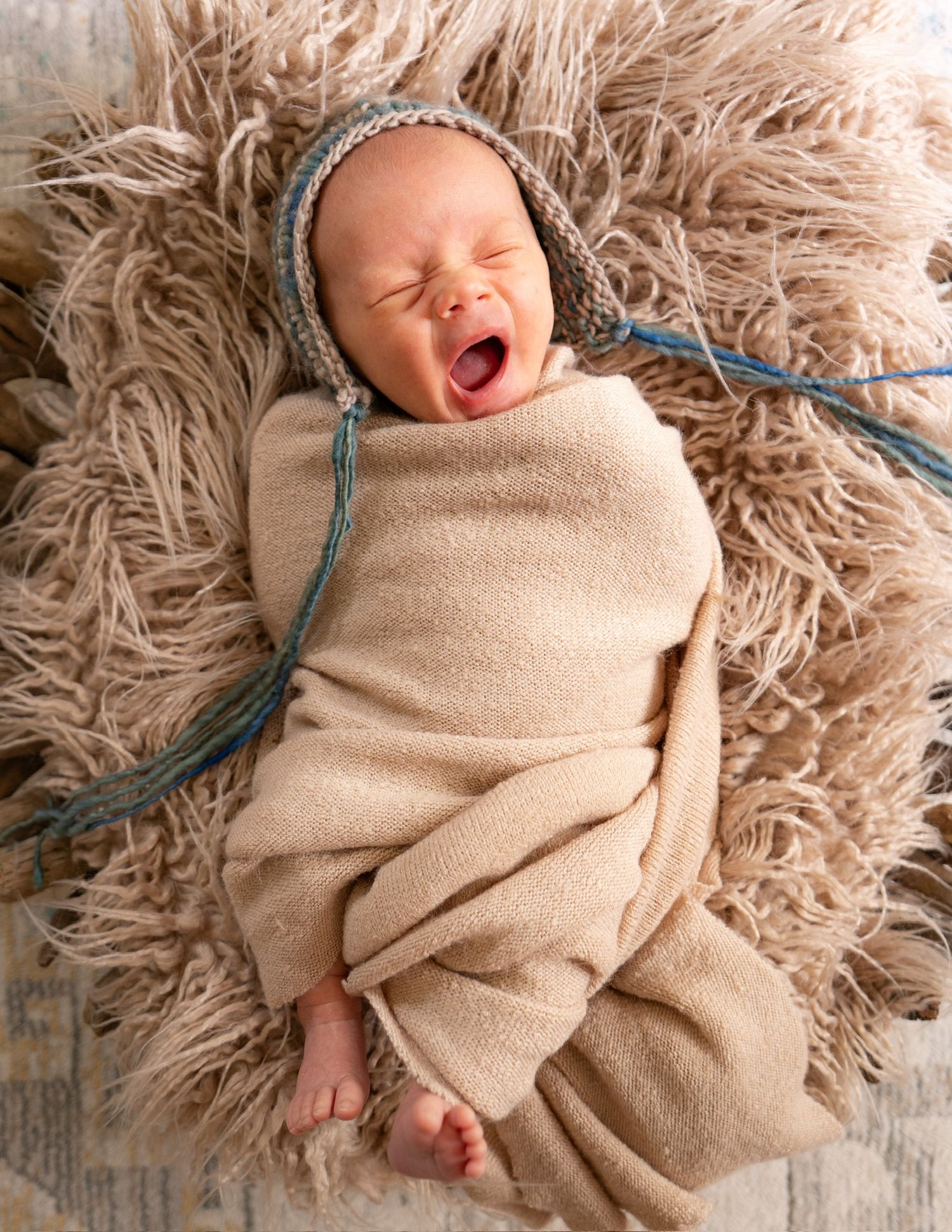 Newborn baby wrapped in beige blanket lying on fluffy rug in progressive stages of expression.