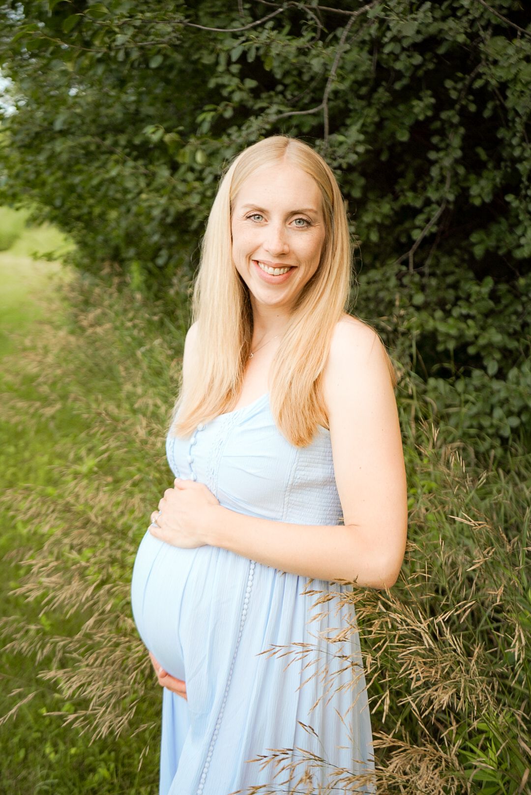 Maternity portrait of expectant mother in light blue dress standing outdoors against green foliage.
