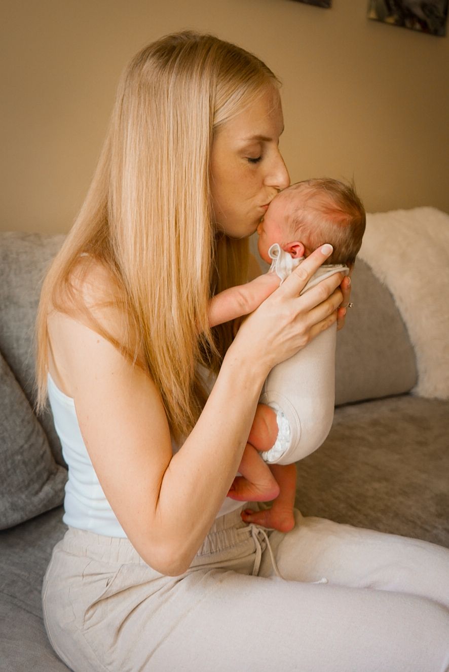 Mother kisses her baby's tiny feet while sitting on a gray couch in a warm-toned room.