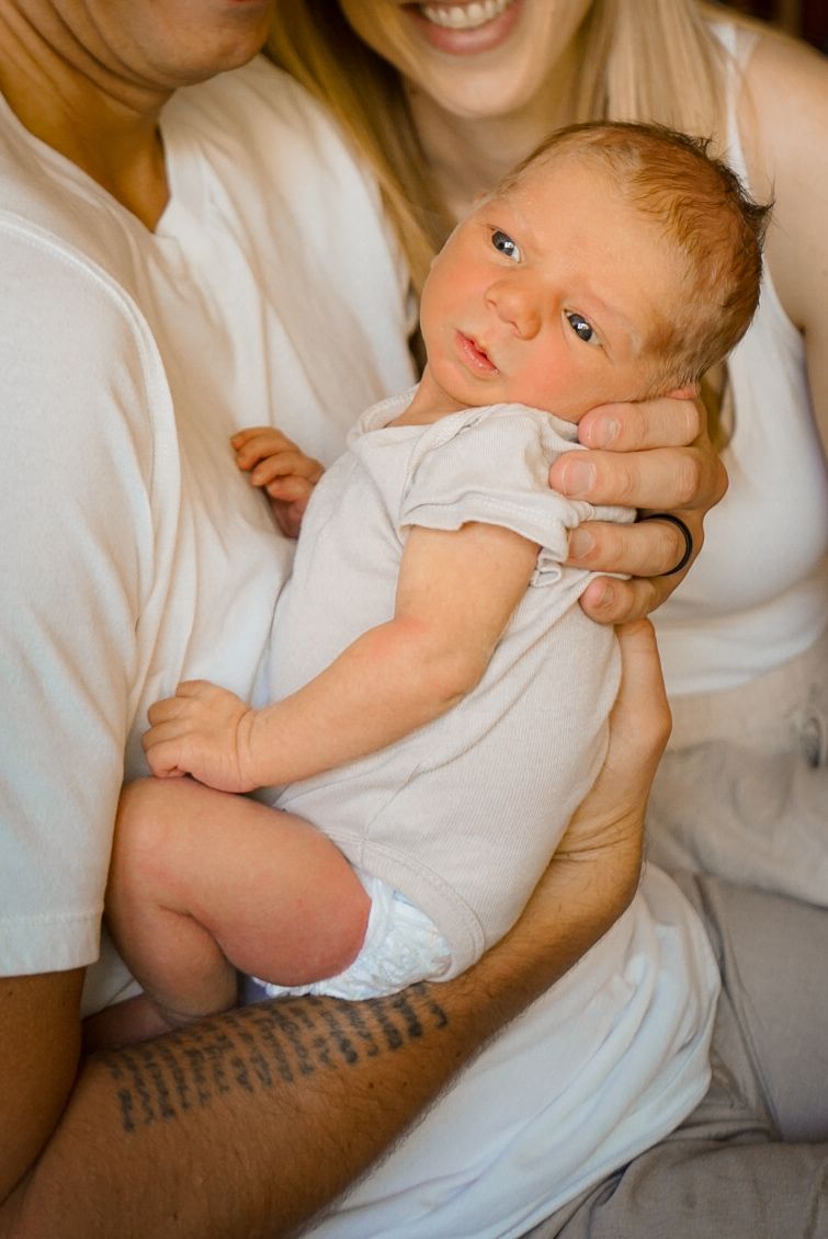 Baby with bright blue eyes wearing white onesie being held and admired by parents.