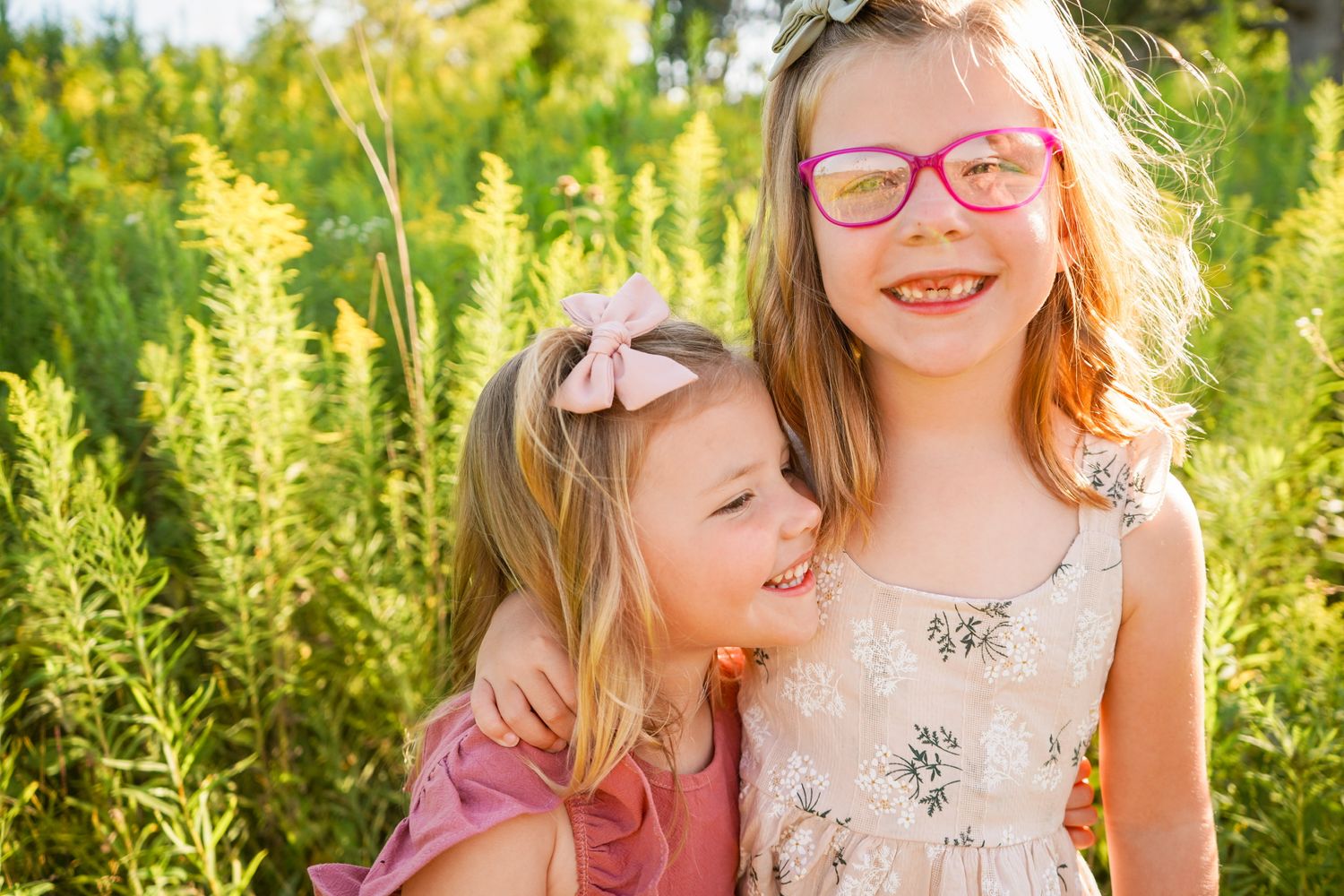 Two children in floral dresses stand together outdoors among green foliage.