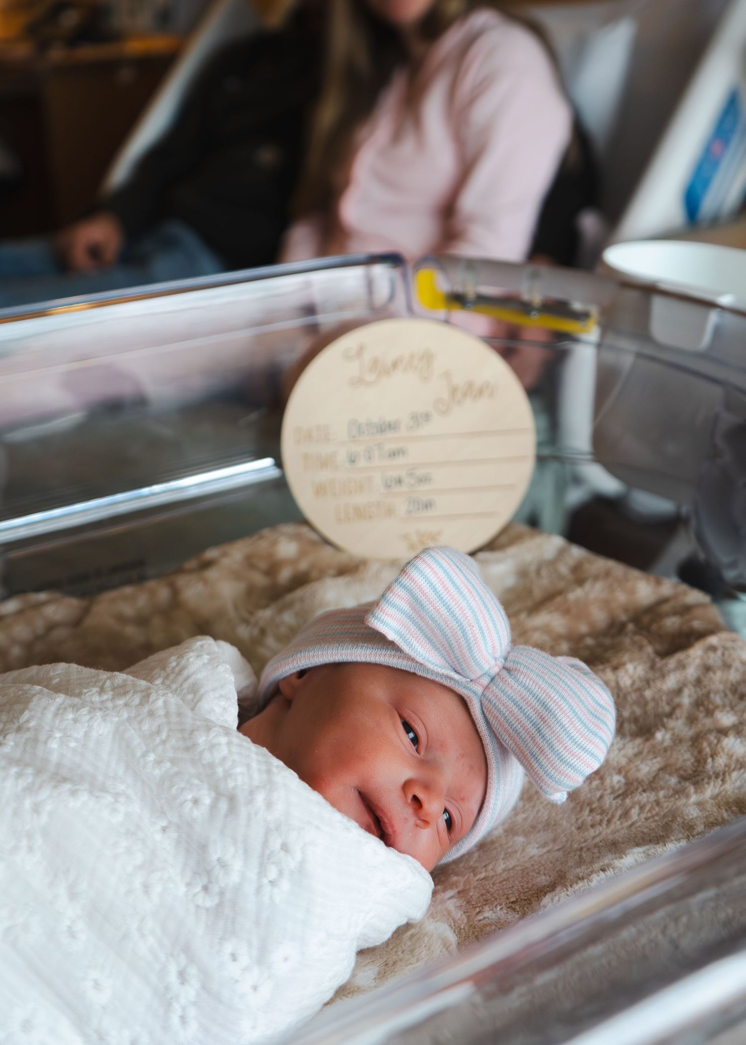 A newborn baby wearing a white bow cap rests peacefully in a hospital bassinet.