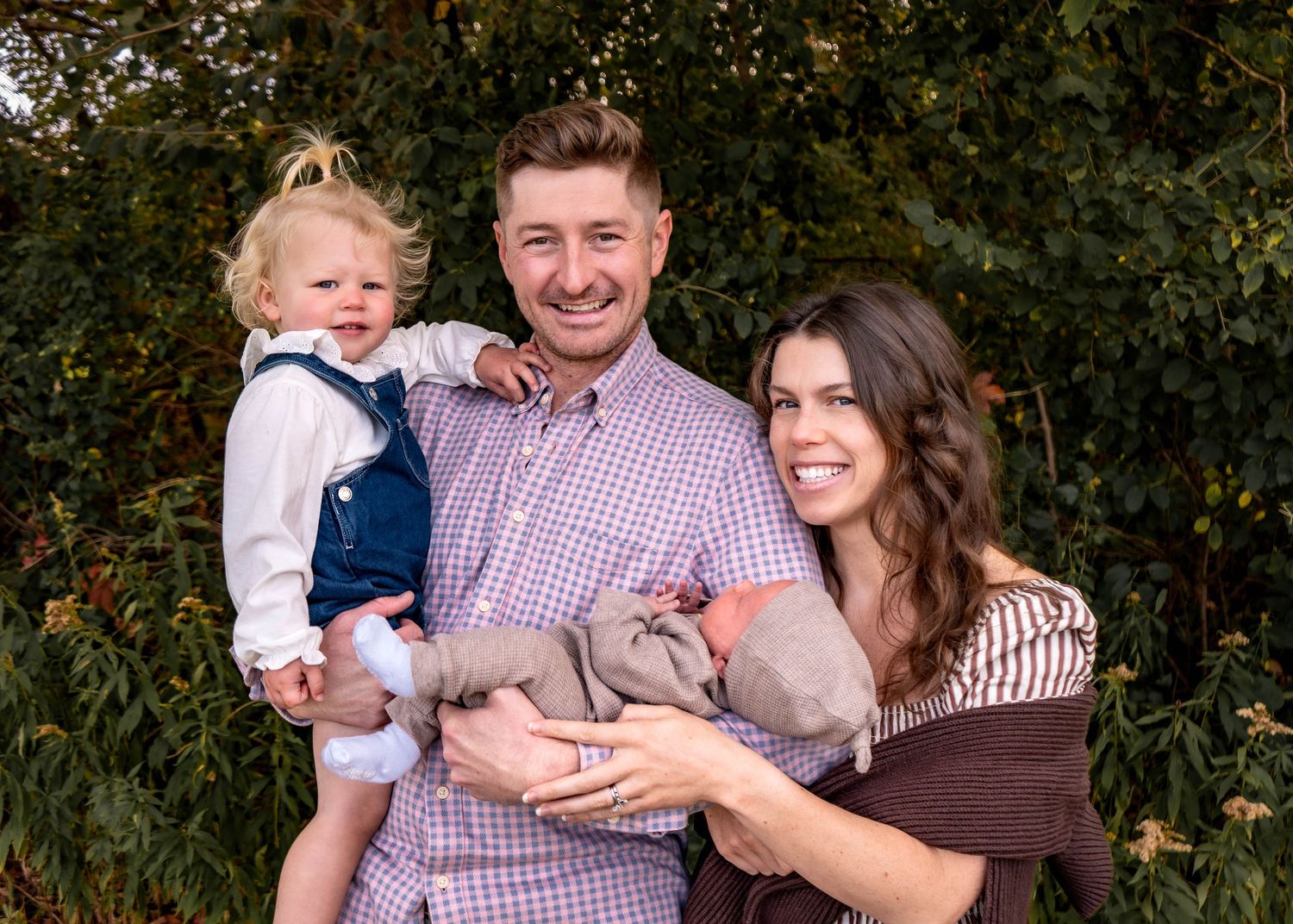 Family portrait in coordinating outfits standing together against green foliage background.