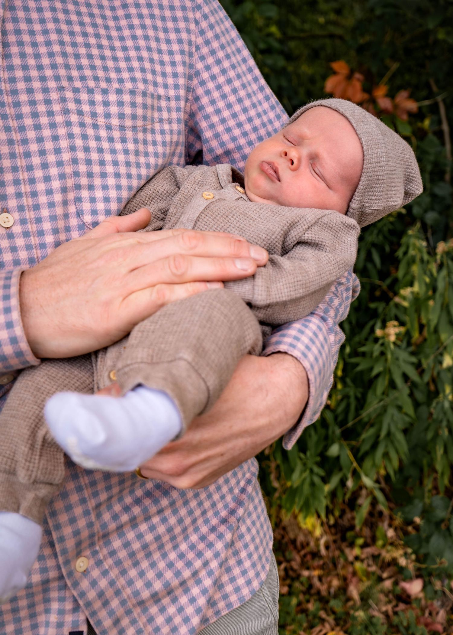 A baby wearing a gray sweater is being held against a checkered shirt with greenery in the background.