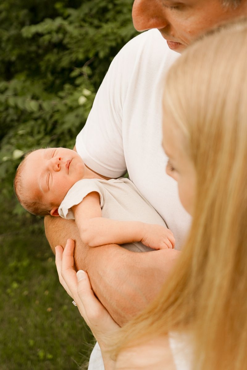 Natural light photography of family embracing their newborn baby outdoors in summer setting.