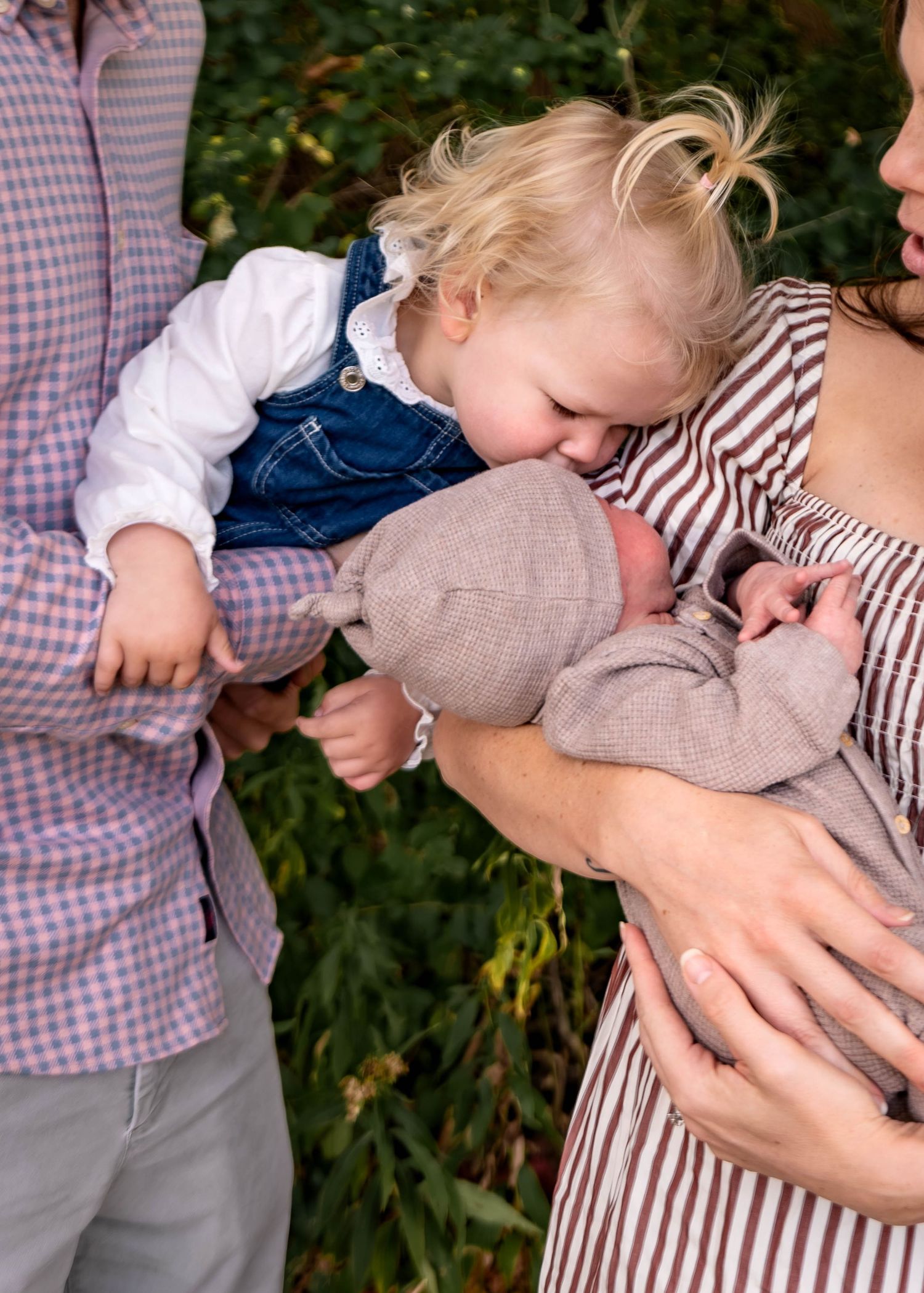 A toddler in denim overalls leaning down to kiss a newborn baby held in someone's arms.
