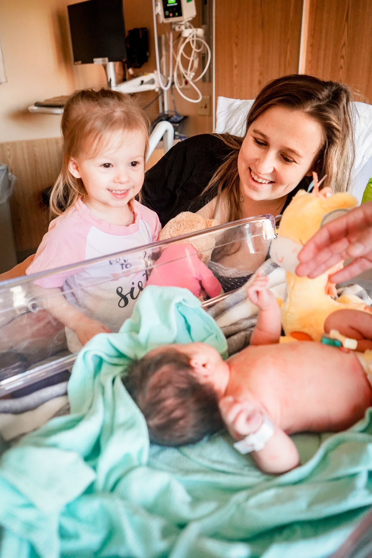 Family members gather around a newborn in a hospital bassinet with stuffed animals and mint green blankets.