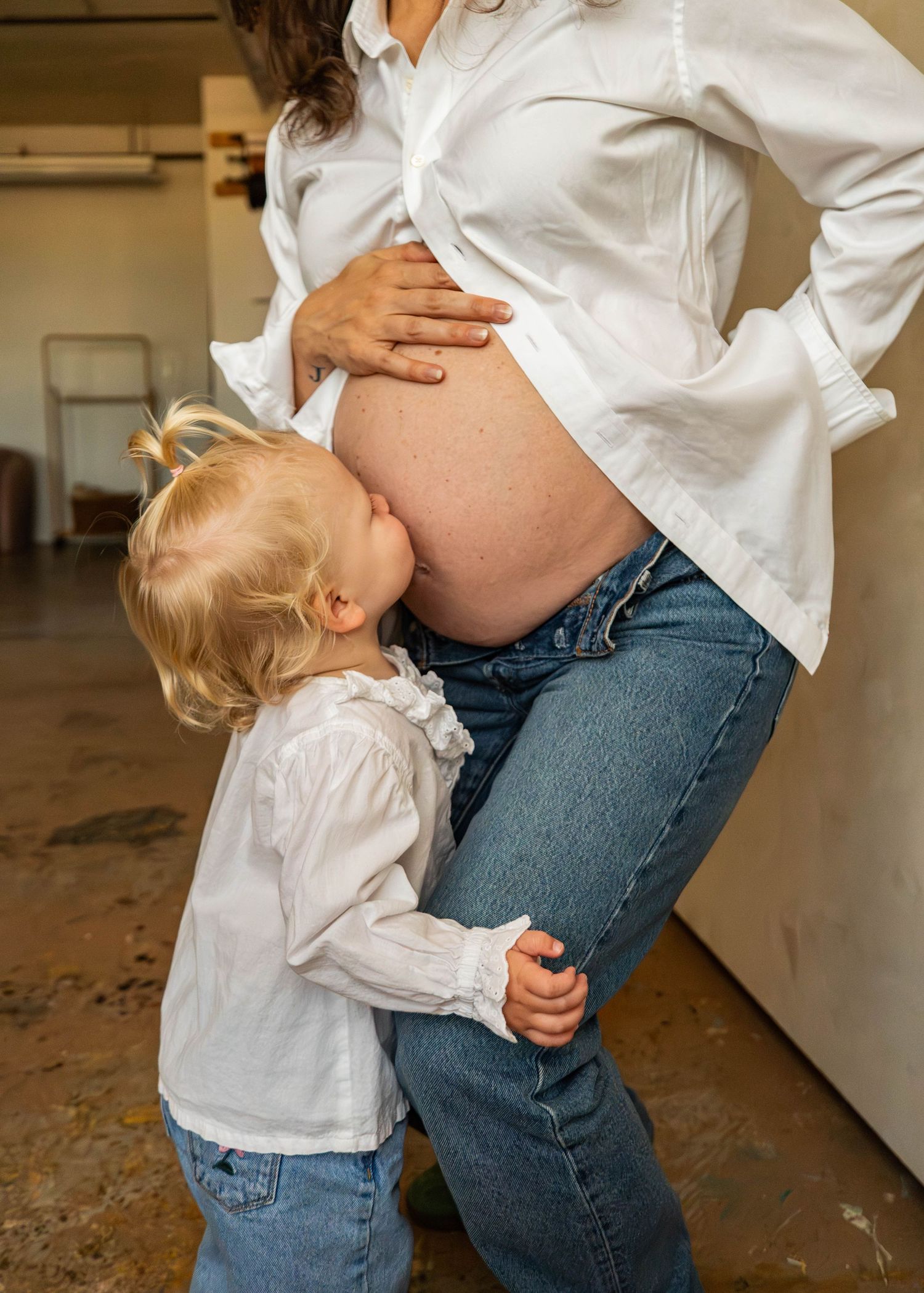 Toddler in white shirt hugging pregnant belly while standing against beige wall.