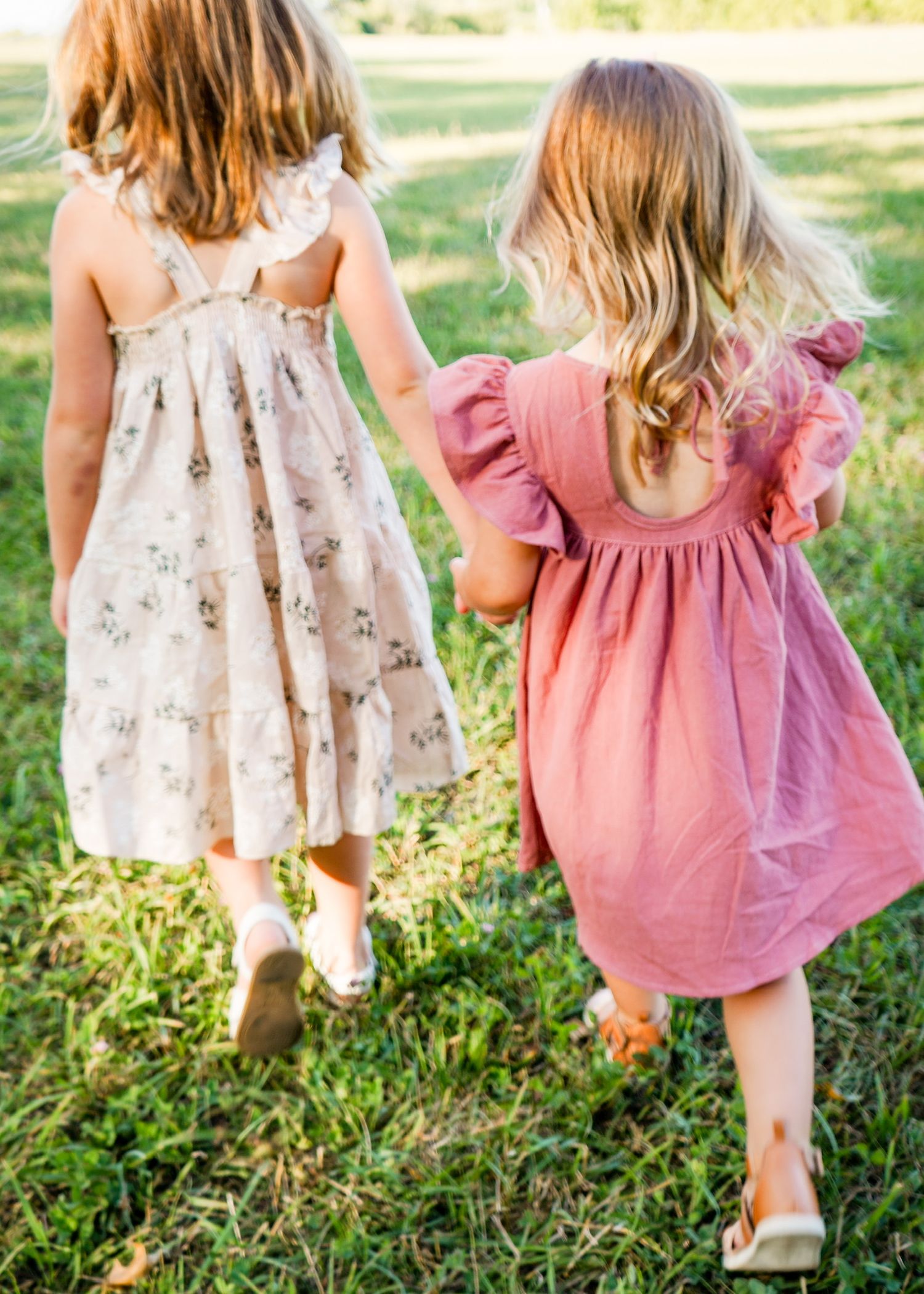Young children in flowing dresses walk through sunlit grass during golden hour.