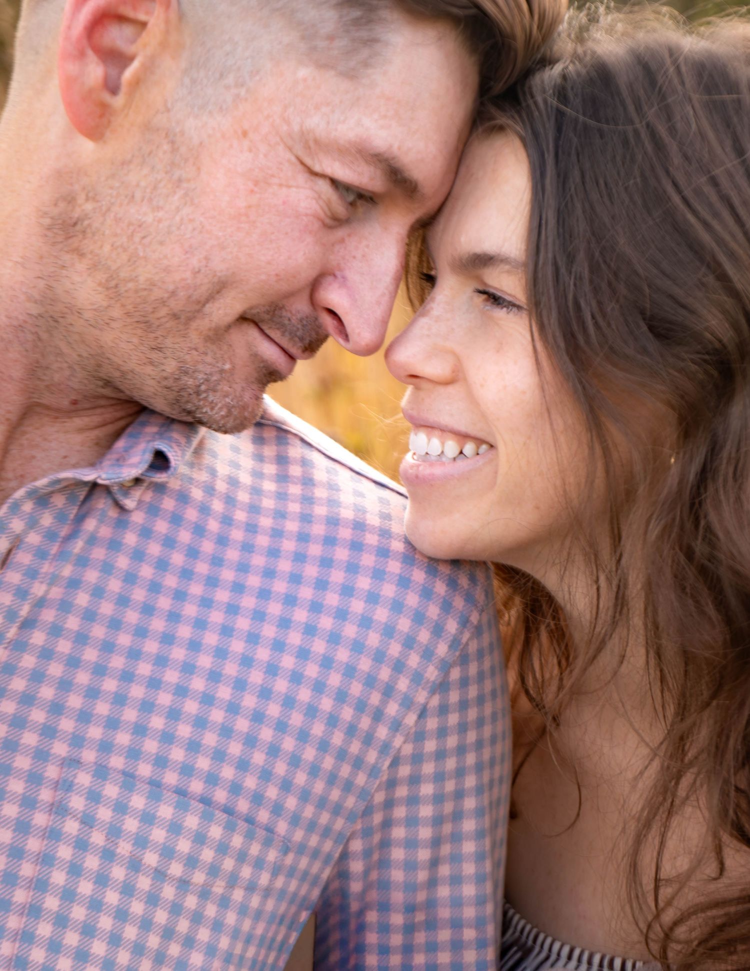 A close-up intimate moment between a couple sharing a tender smile against a bright background.