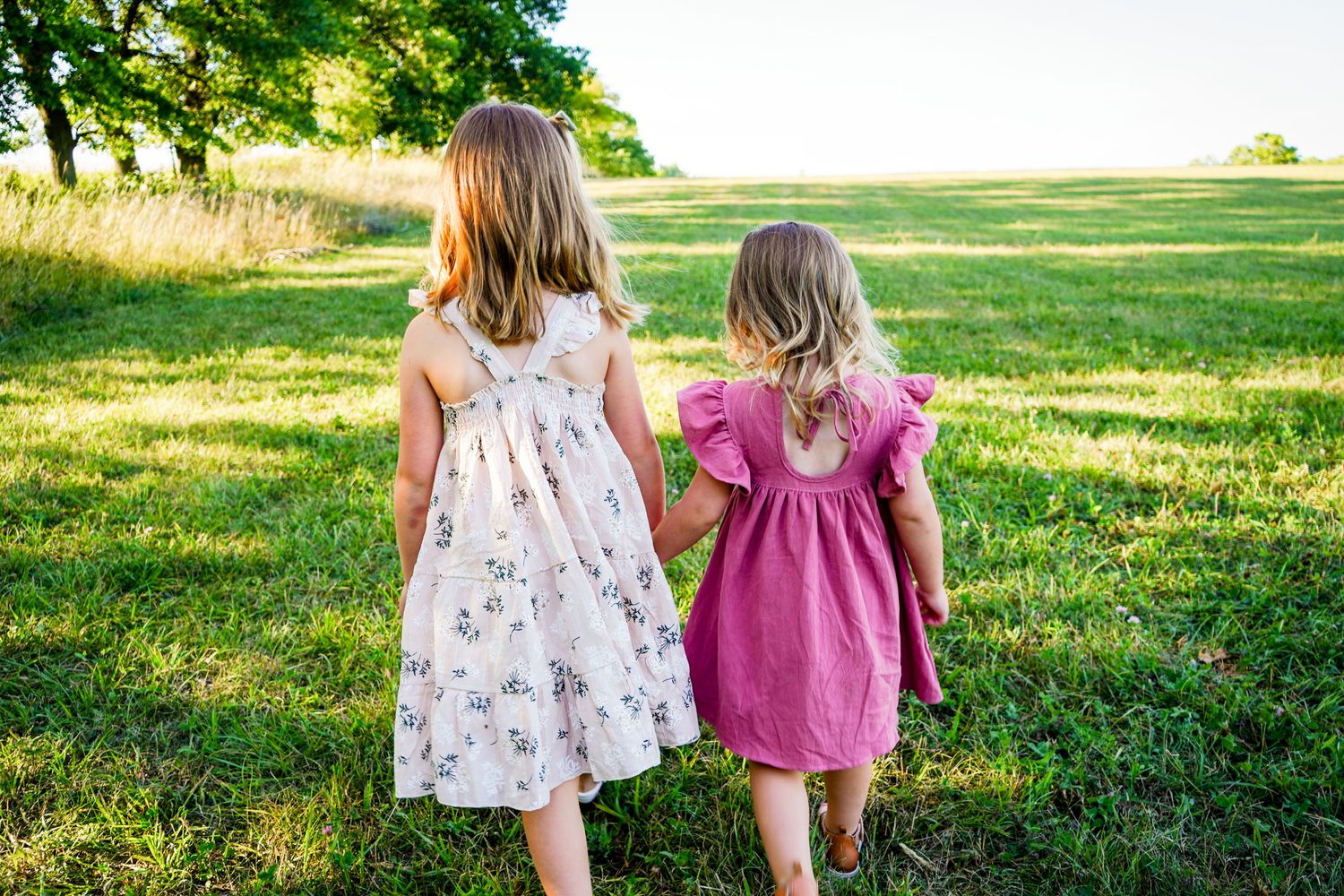 Two young figures in summer dresses walk hand-in-hand through a sunny grassy field.