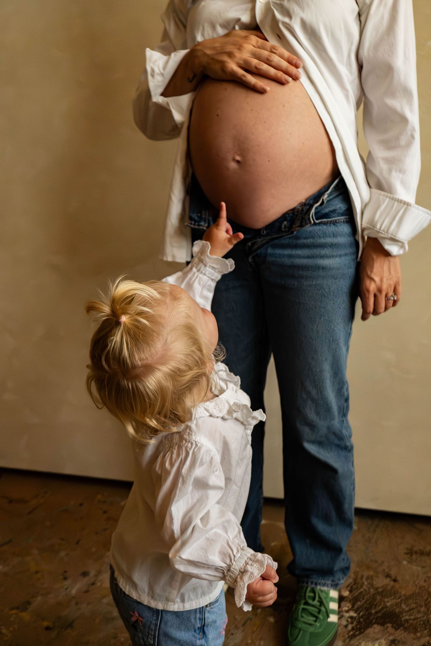 A toddler in white shirt reaches up to touch a pregnant belly wearing jeans and white button-down shirt.