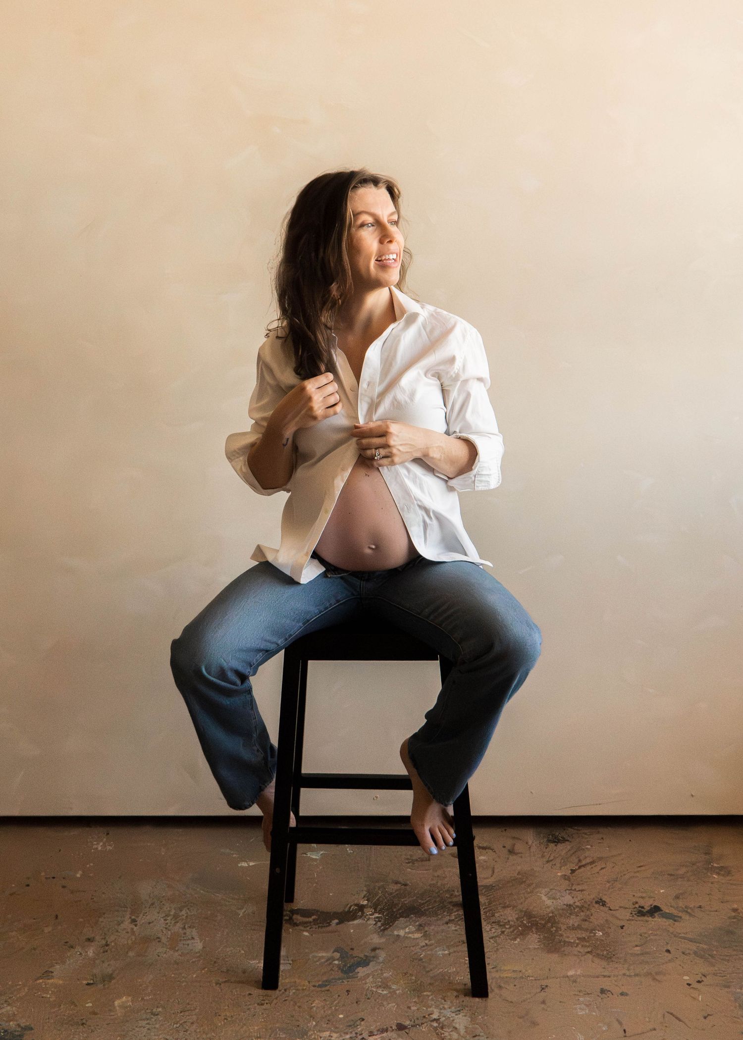 Pregnant woman in white shirt and jeans sits on dark stool against beige wall.