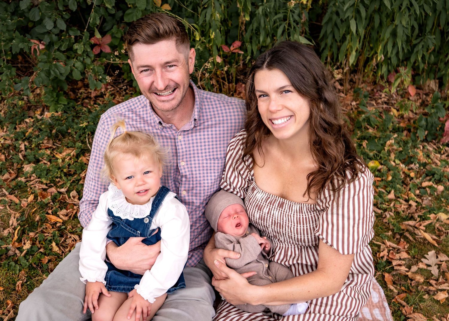 A family portrait outdoors with a toddler in overalls and a newborn baby against a leafy background.