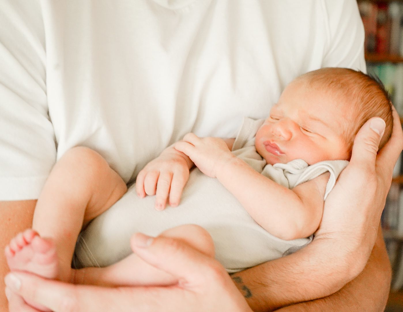 A sleeping baby in light gray clothing nestled securely in caring arms wearing white.