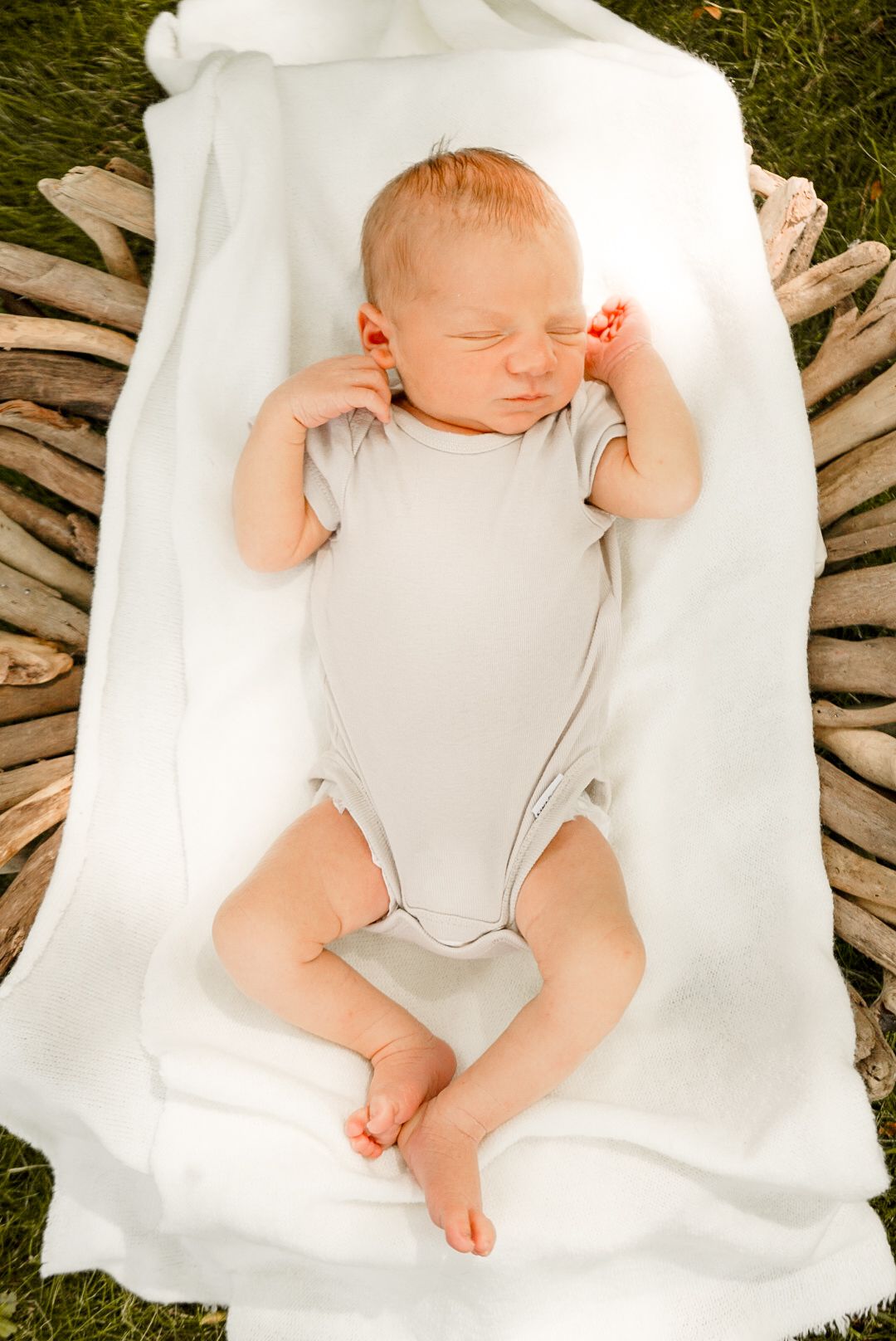 A peaceful sleeping infant wearing grey clothing rests on white fabric against a natural backdrop.