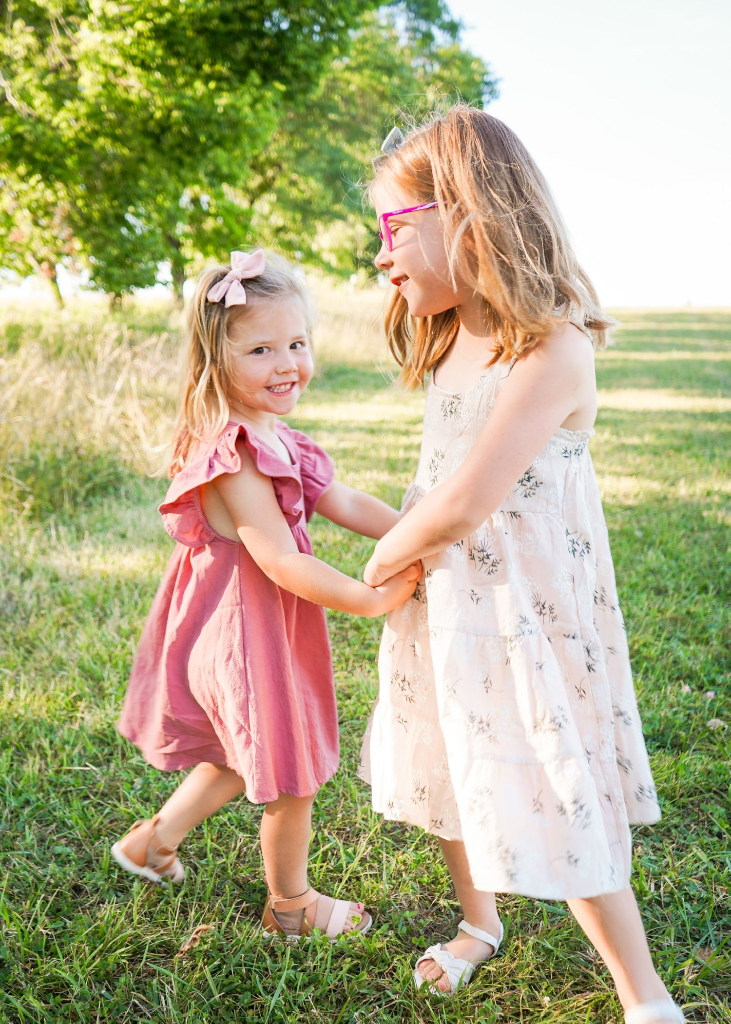 Two children in summer dresses playing and dancing together in a grassy field on a sunny day.