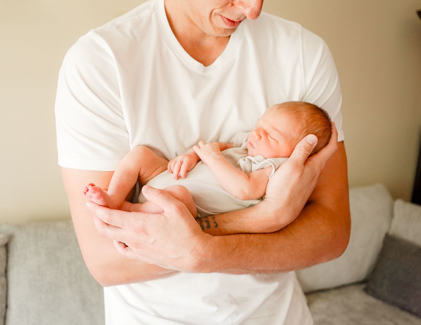 A sleeping newborn baby in a light colored onesie being cradled in arms wearing a white t-shirt.