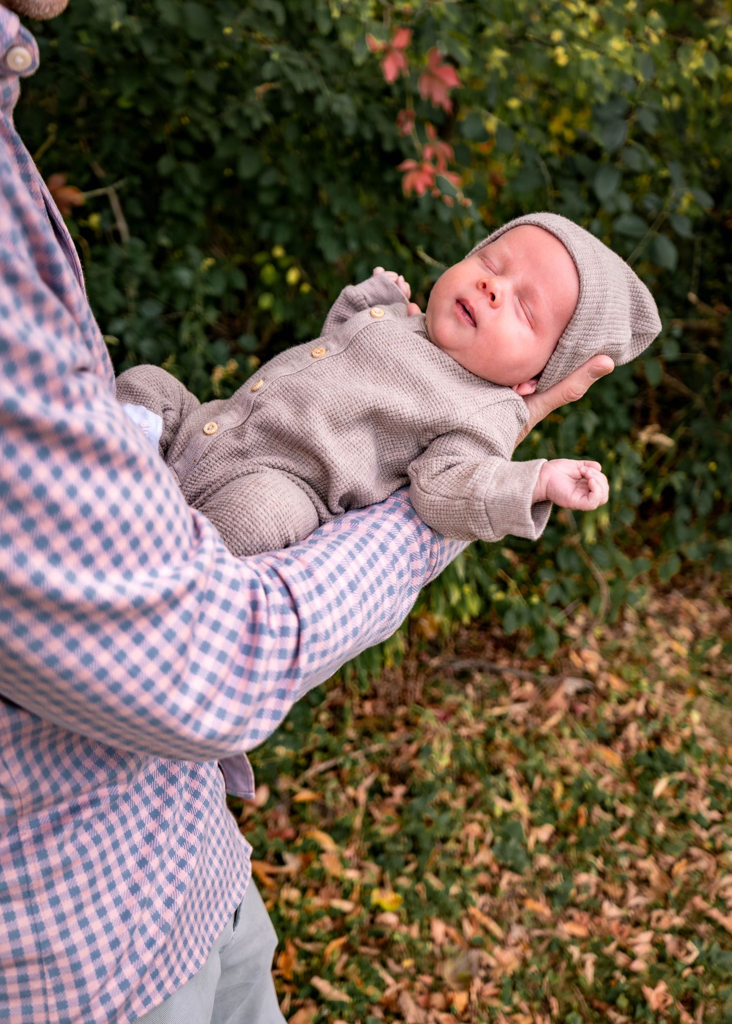 Baby wearing a soft gray sweater being held against autumn foliage background.
