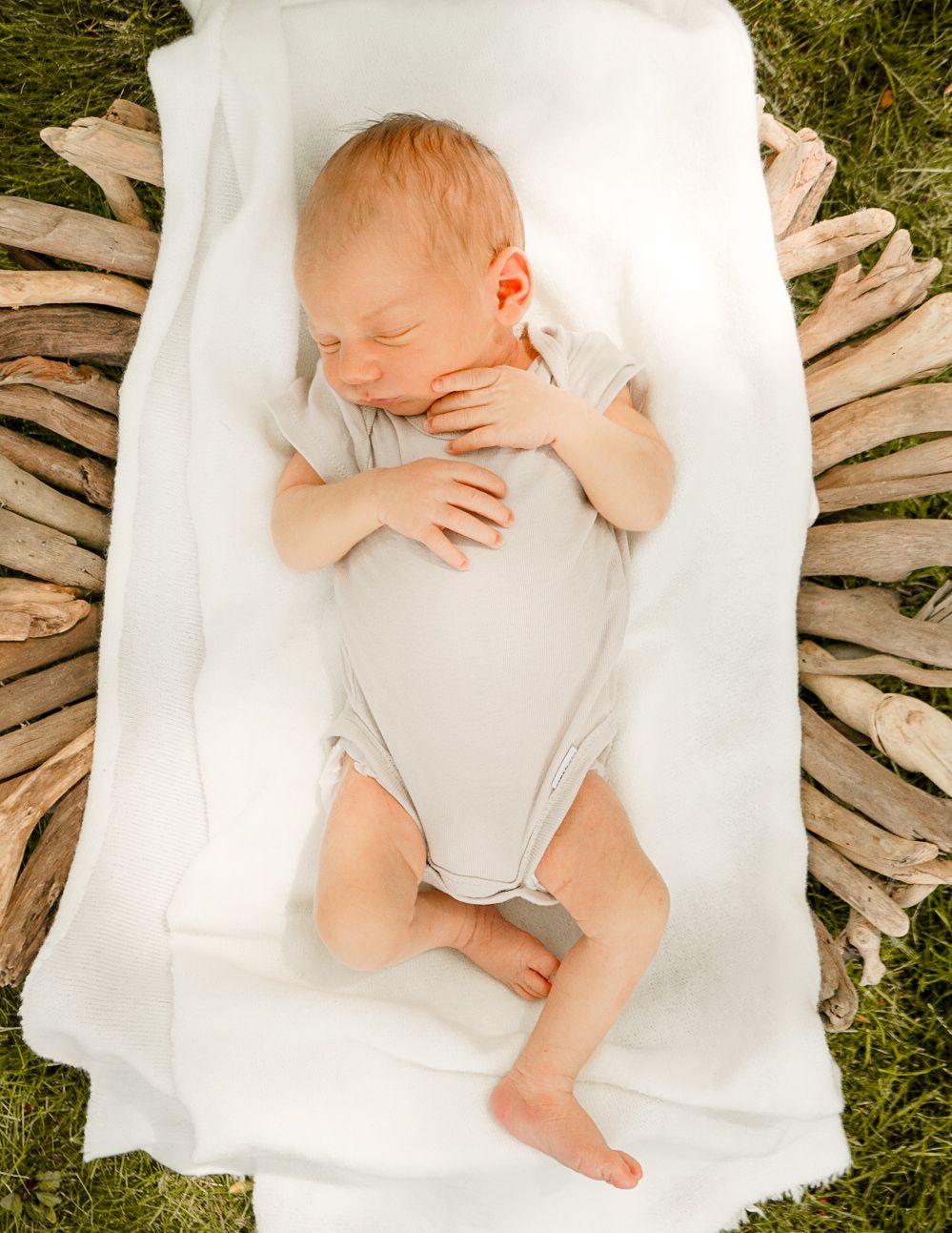 Peaceful infant wearing gray onesie sleeps on crisp white linens against natural woven backdrop.