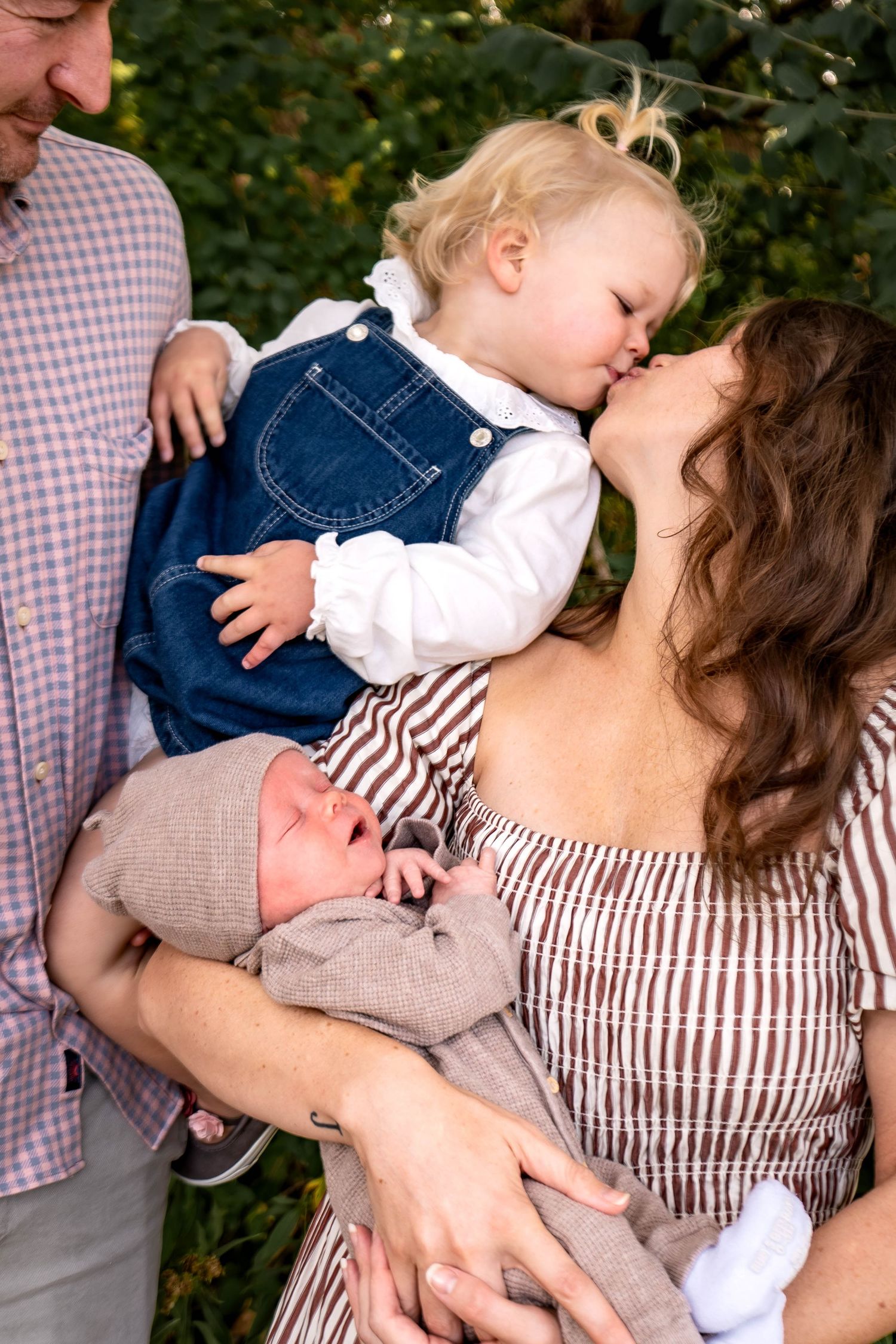 Tender family moment outdoors with toddler in denim overalls and baby in grey sweater being held close.