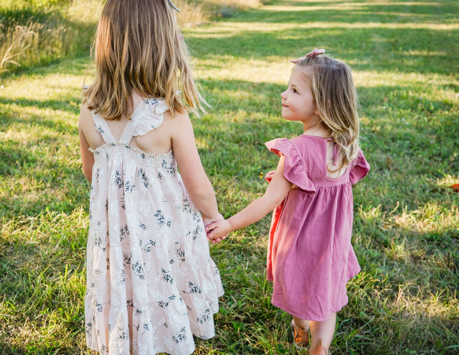 Two kids in light floral and pink dresses playing together outdoors on grass.
