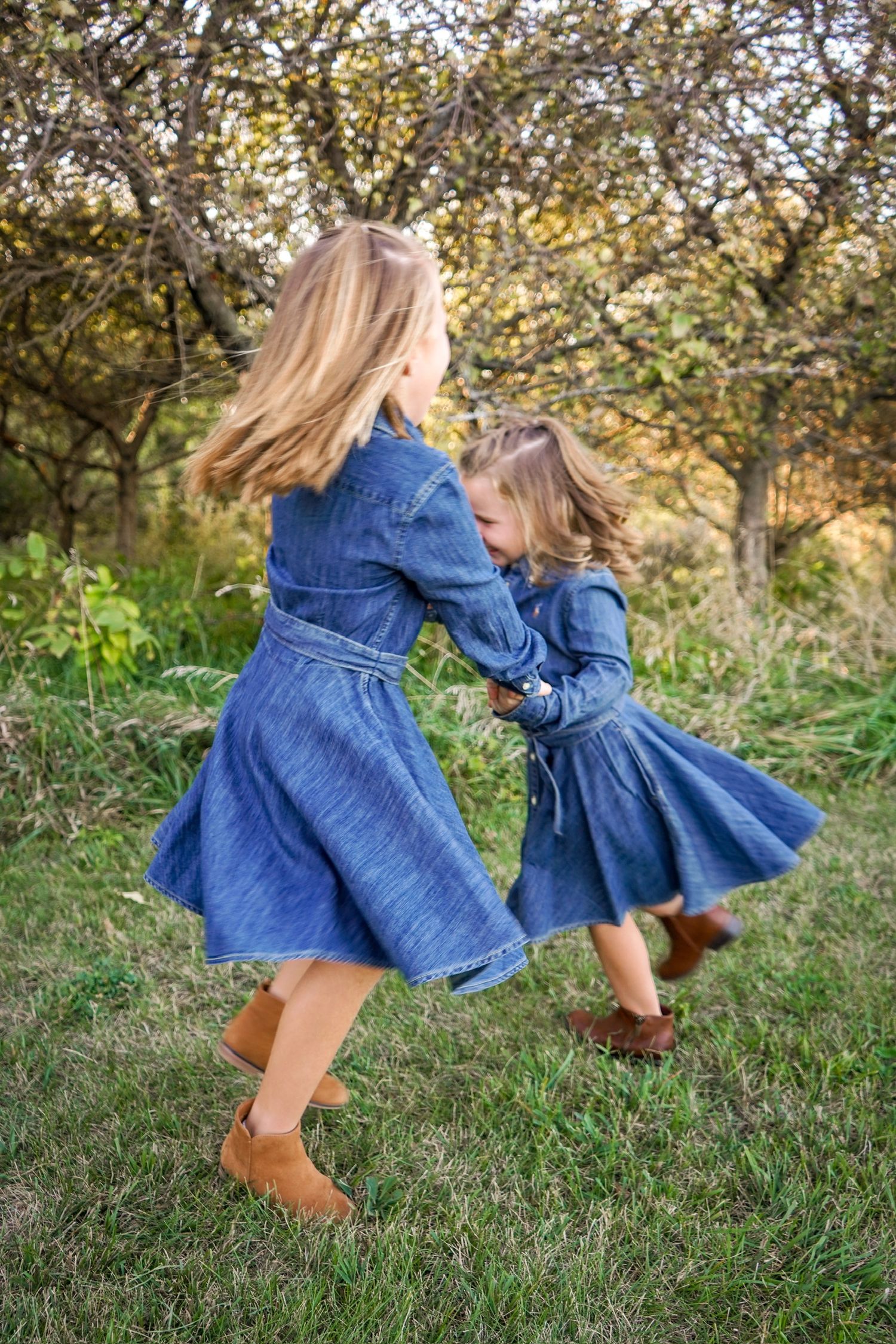 Children in denim dresses twirling and playing in a grassy field during golden hour.