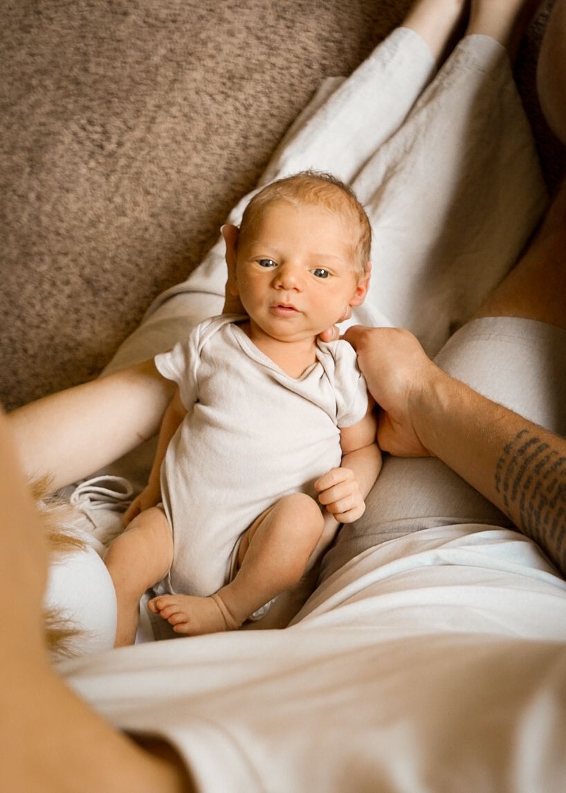 An infant wearing a white onesie rests on light-colored bedding in warm natural light.