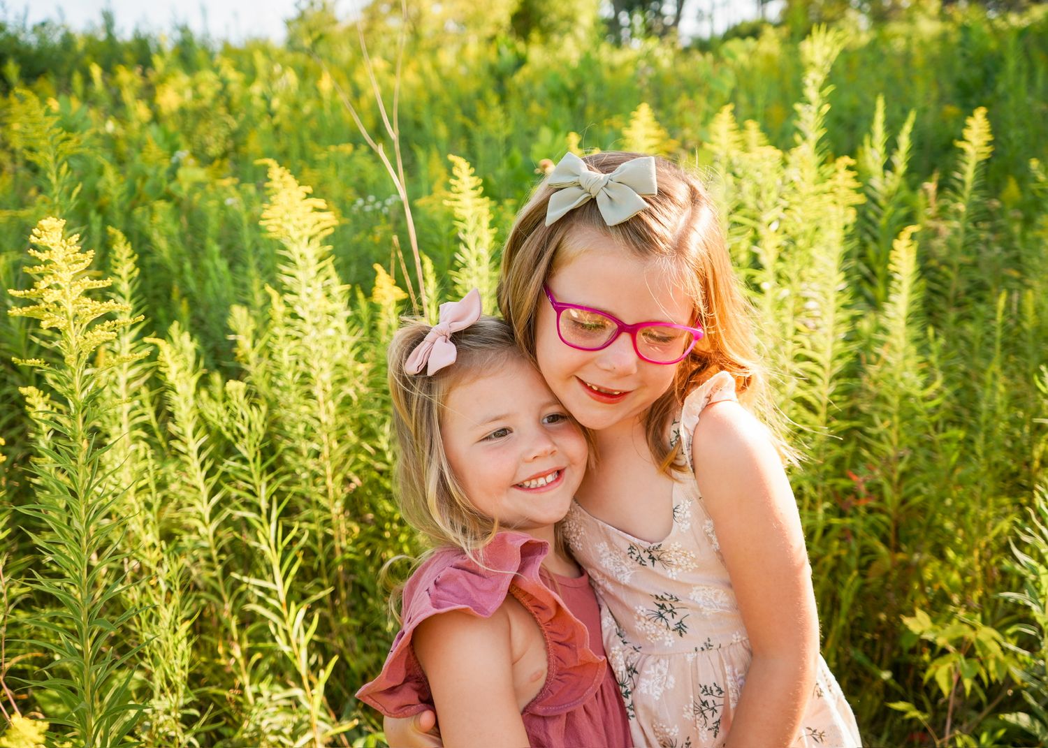 Two children embracing while sitting among lush green foliage on sunny day.
