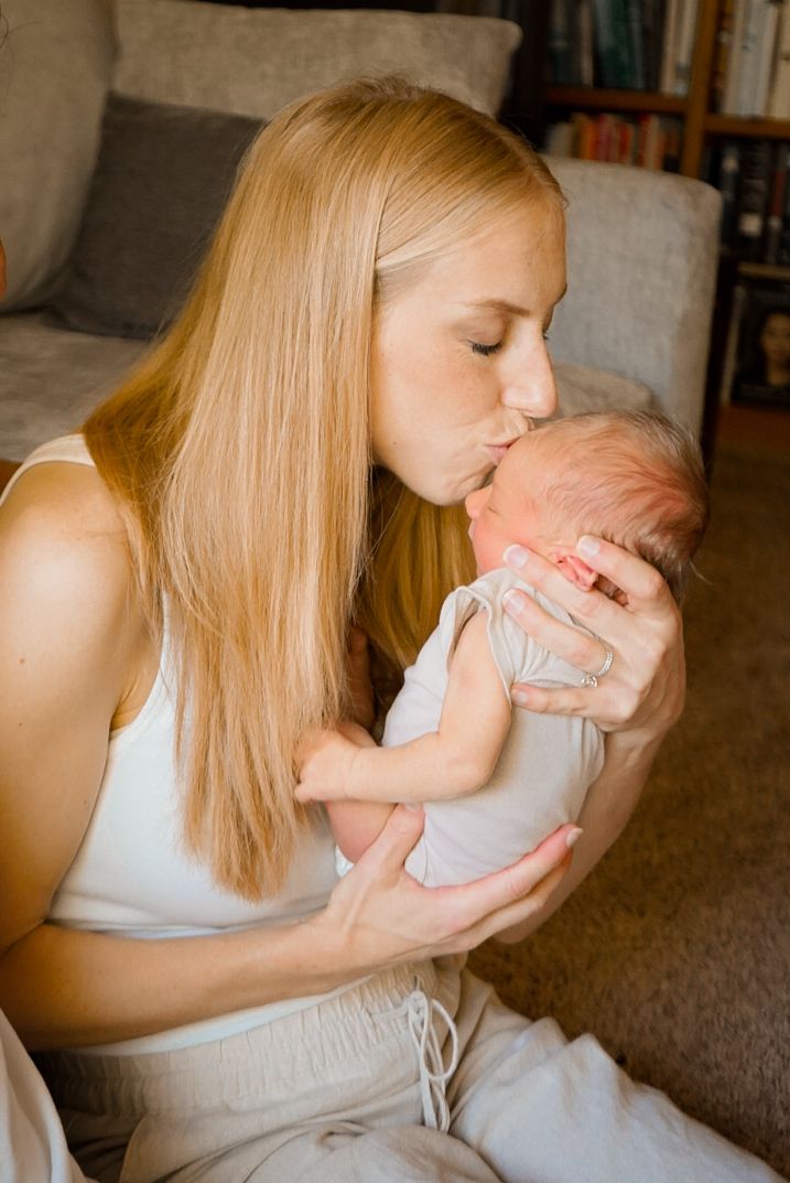 Woman in white holding and kissing a newborn baby in a soft-lit room.