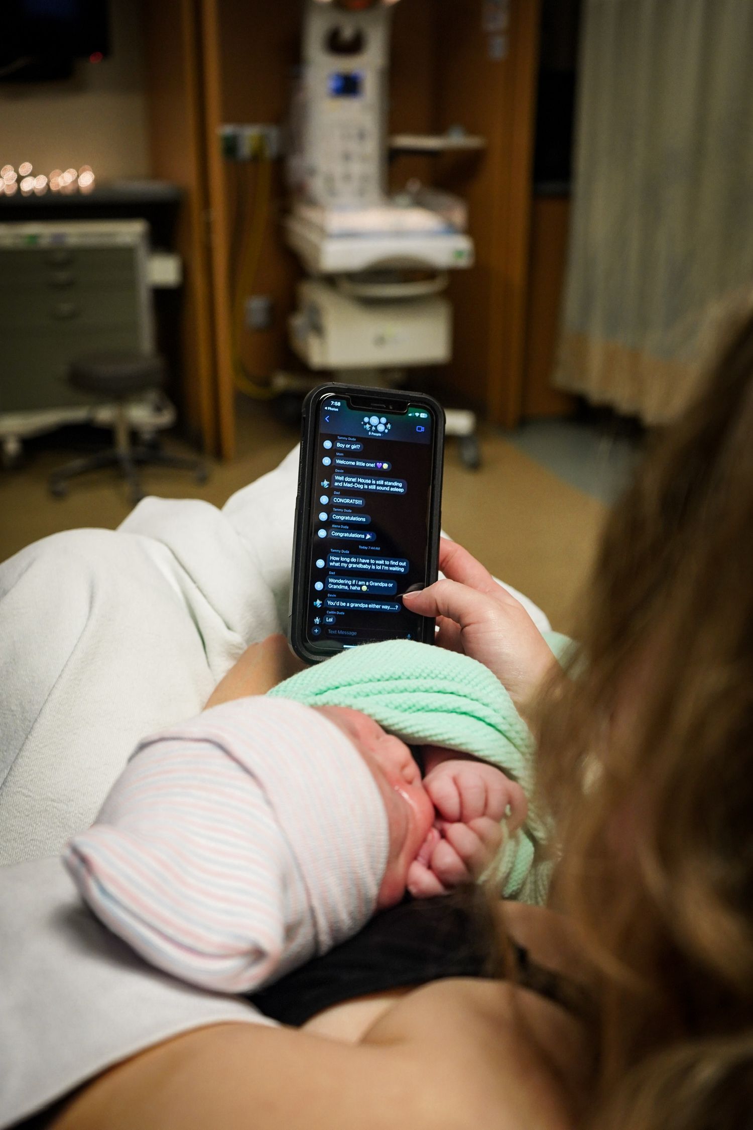 Parent using smartphone while resting in hospital bed with newborn.