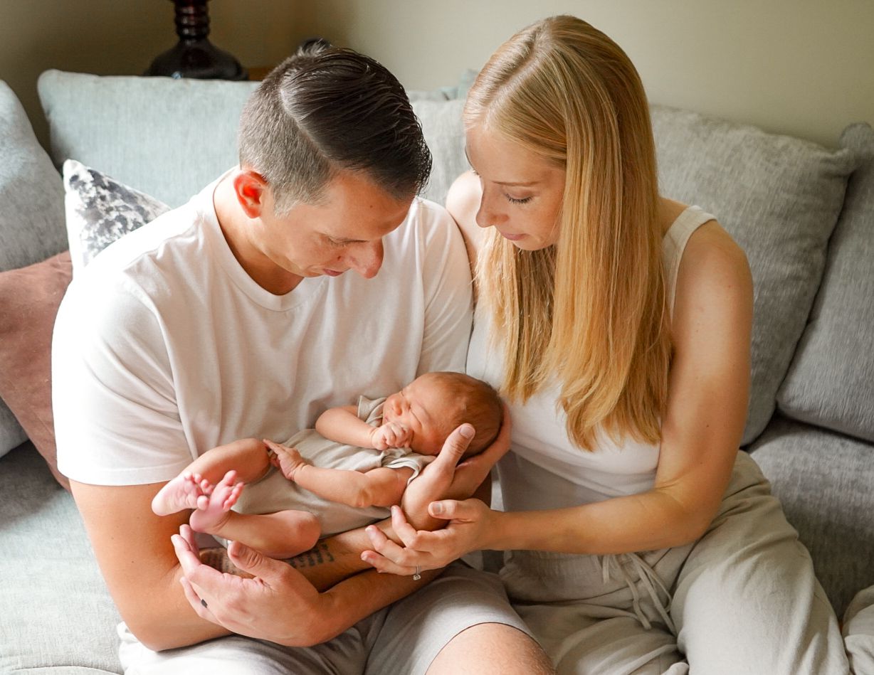 A couple sits together on a gray couch examining their newborn baby in an intimate setting.