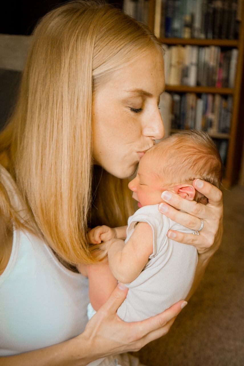 Tender moment captured as woman gently kisses newborn baby near bookshelf.
