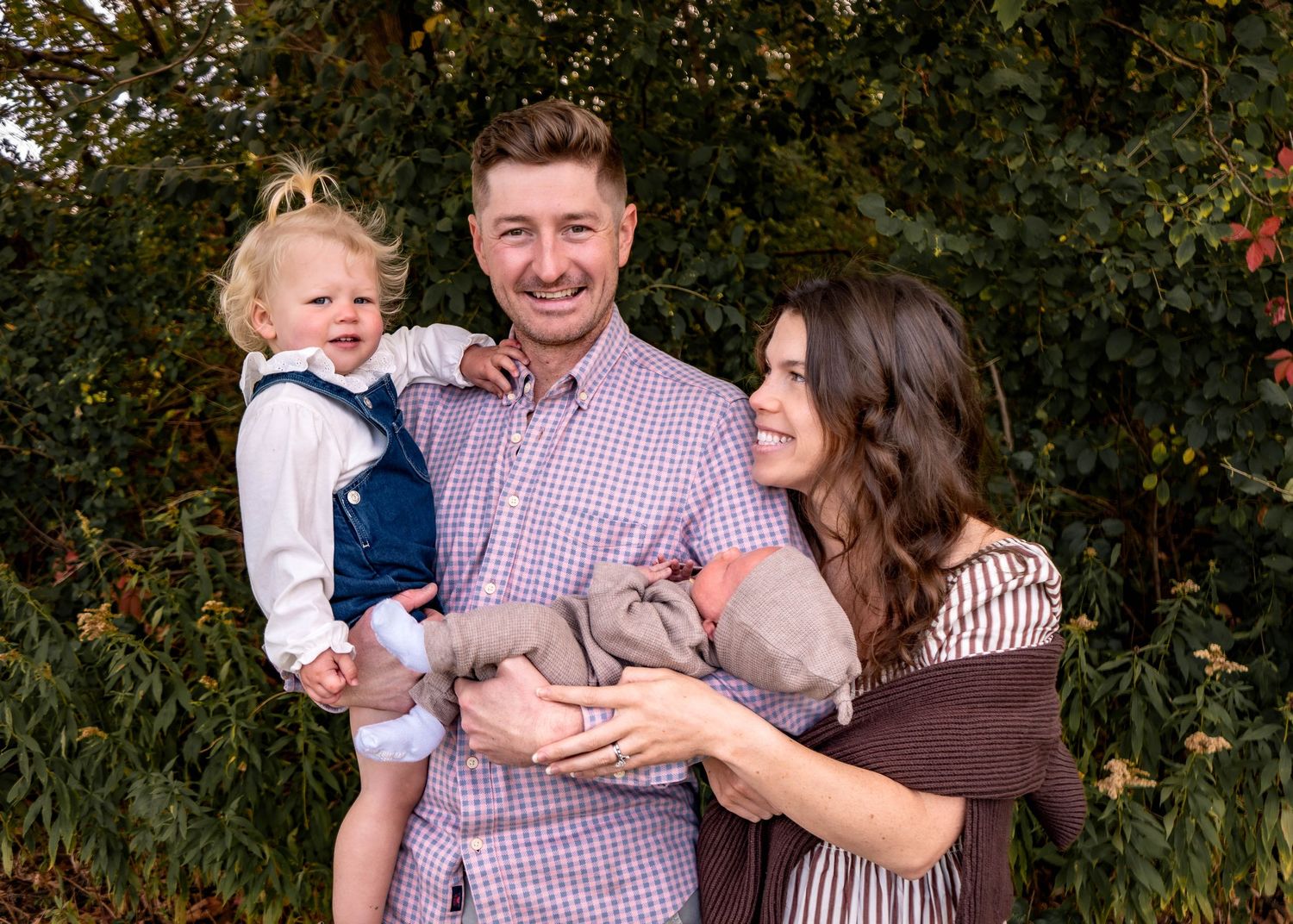 A family embraces outdoors in front of autumn foliage wearing coordinating purple and brown outfits.