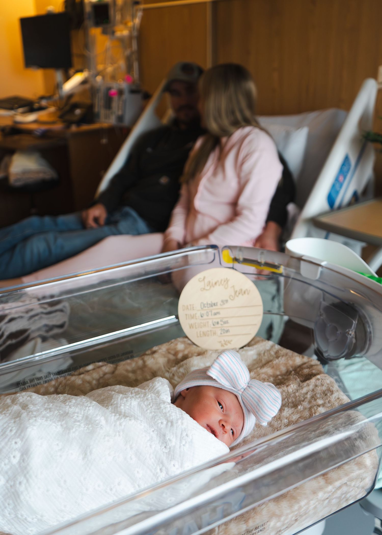 A newborn baby rests in a hospital bassinet with a white cap while people sit in the background.